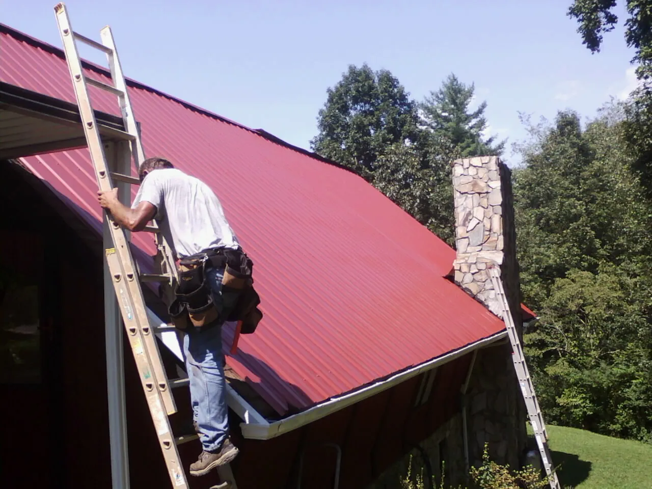 Roofer on a ladder beside a red metal roof and stone chimney — Soffit & Fascia Repair by Roofing Brothers in Maidsville